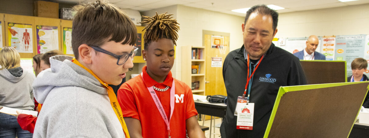 Two male teen students and an adult male look at a poster board