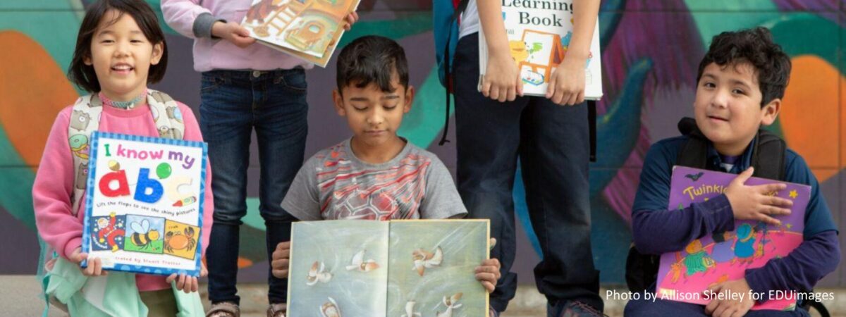 A group of kindergarten and first-grade students pose with their favorite books in front of a school mural.