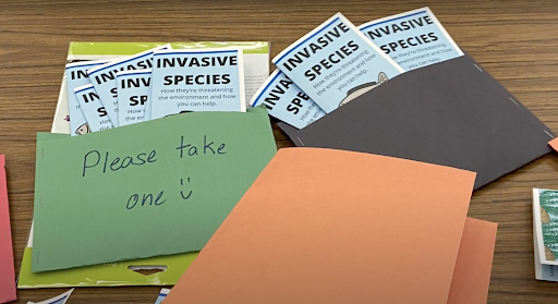 Two folders with Invasive Species brochures sit on a table with a sign reading "take one."