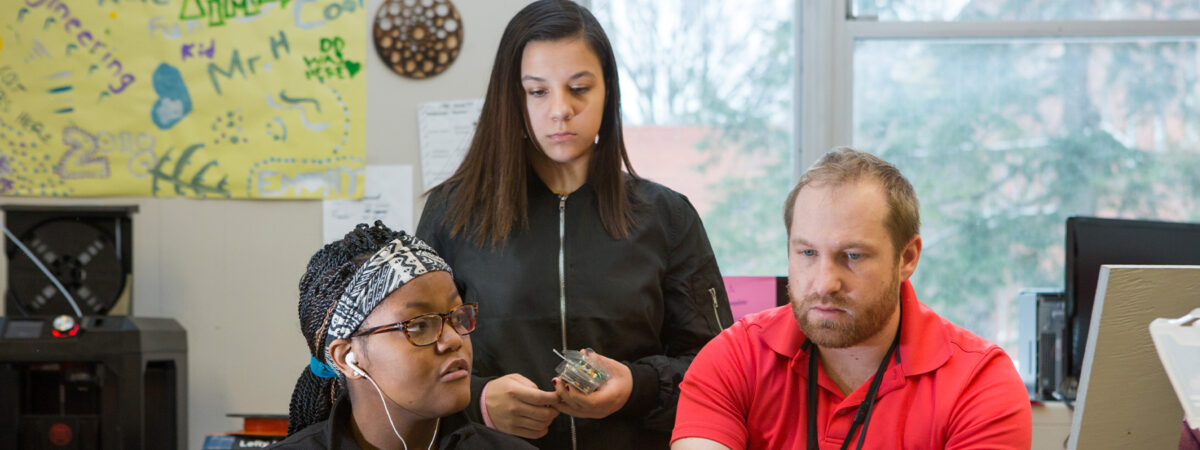 Male teacher helps two female students at computer