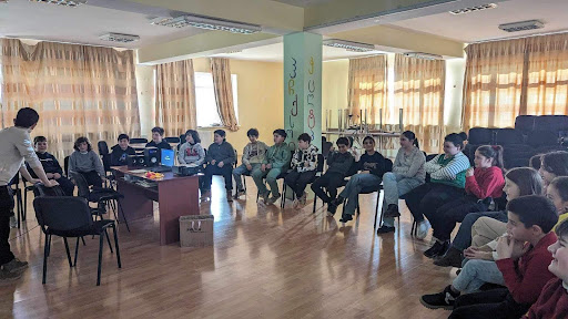 An older student speaks in front of younger students in a school room. 