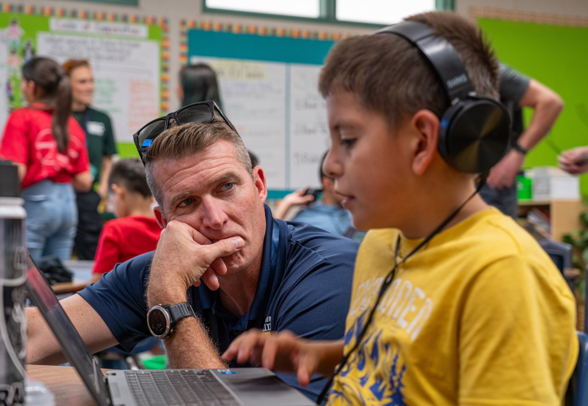 A white male adult speaks with an elementary school aged student wearing headphones at a lap top. 