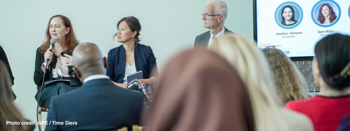 A panel of 3 adults faces an audience. One holds a microphone and speaks.