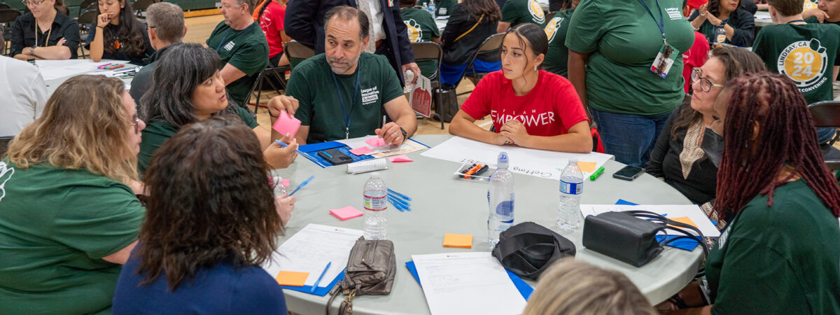 A group of district leaders sit at a table with a student working on a project