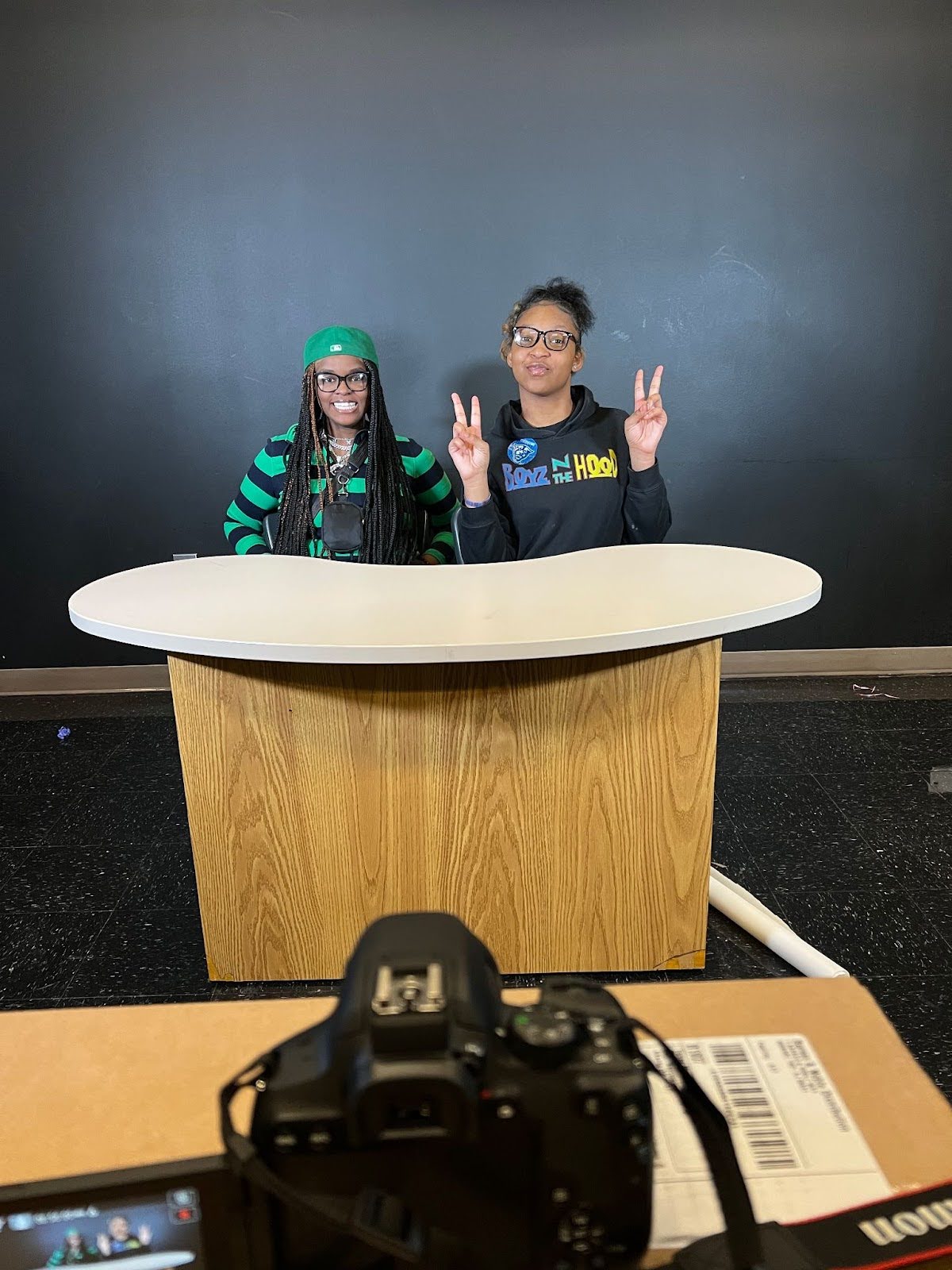 Two students are seated at a news anchor desk in their studio. 