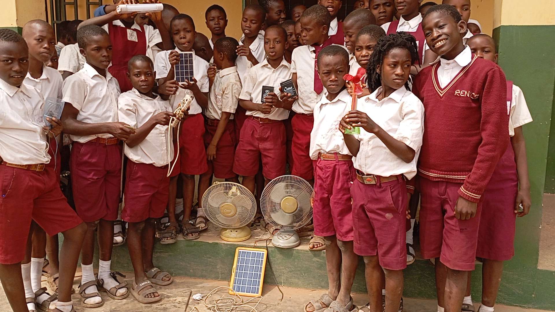 A large group of students hold up their devices in front of their school. 