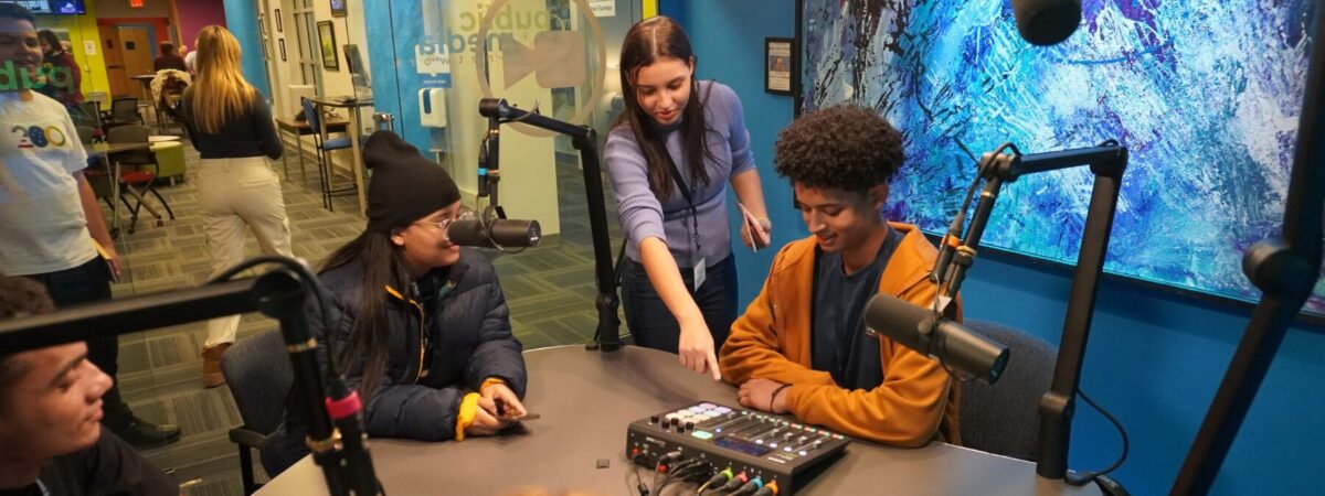 An adult stands behind a student while pointing at a sound mixer. Students are seated at a table with a mixer and microphone.