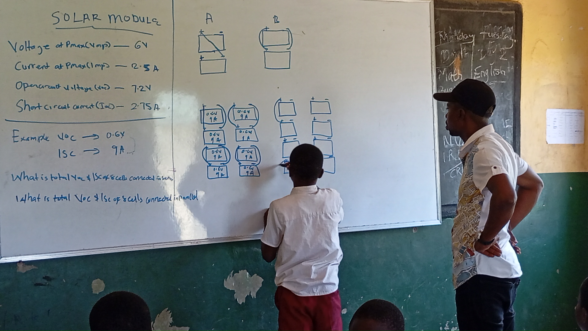 A student writes on a whiteboard while an adult reviews from behind the student. 