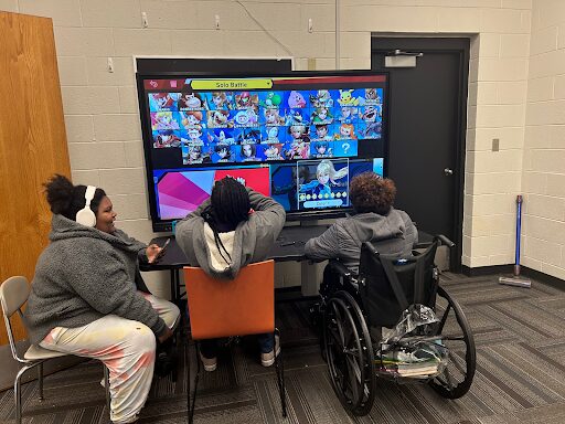 Three students sit facing a large television screen. 