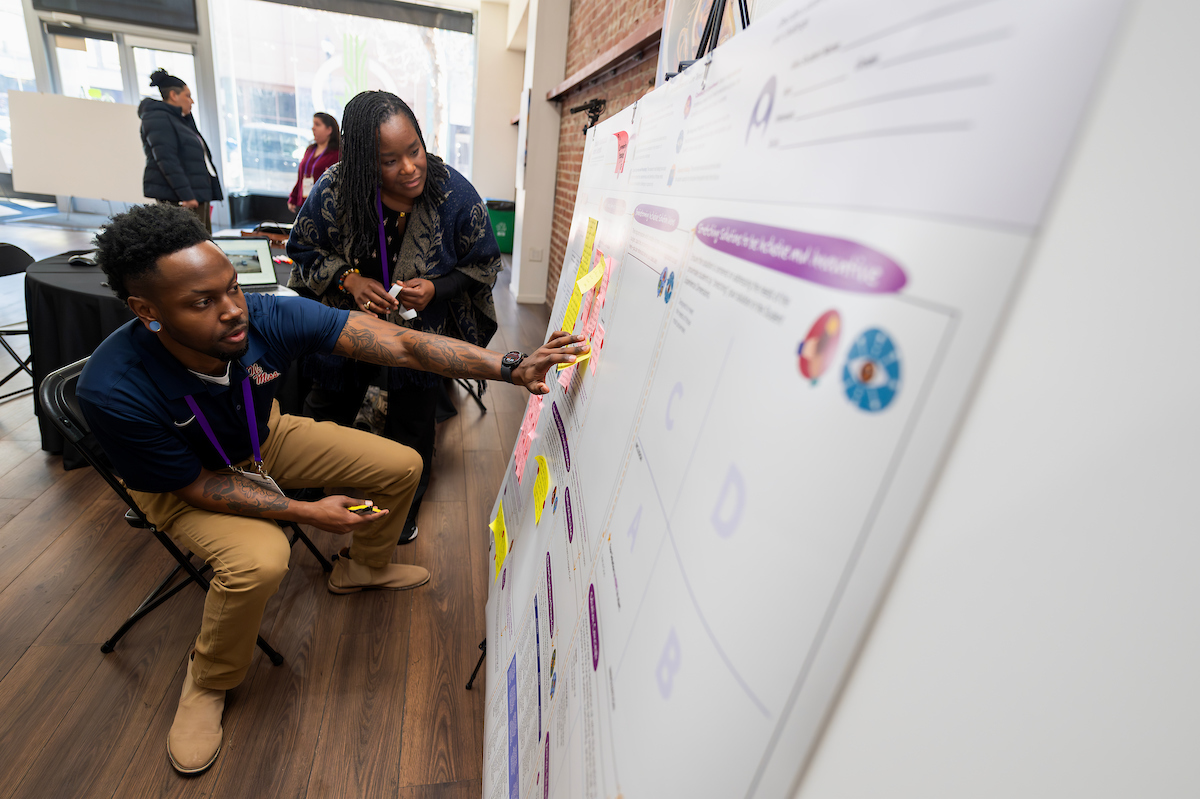 A woman and a young man peer over a workshop poster together, placing sticky notes on its surface.