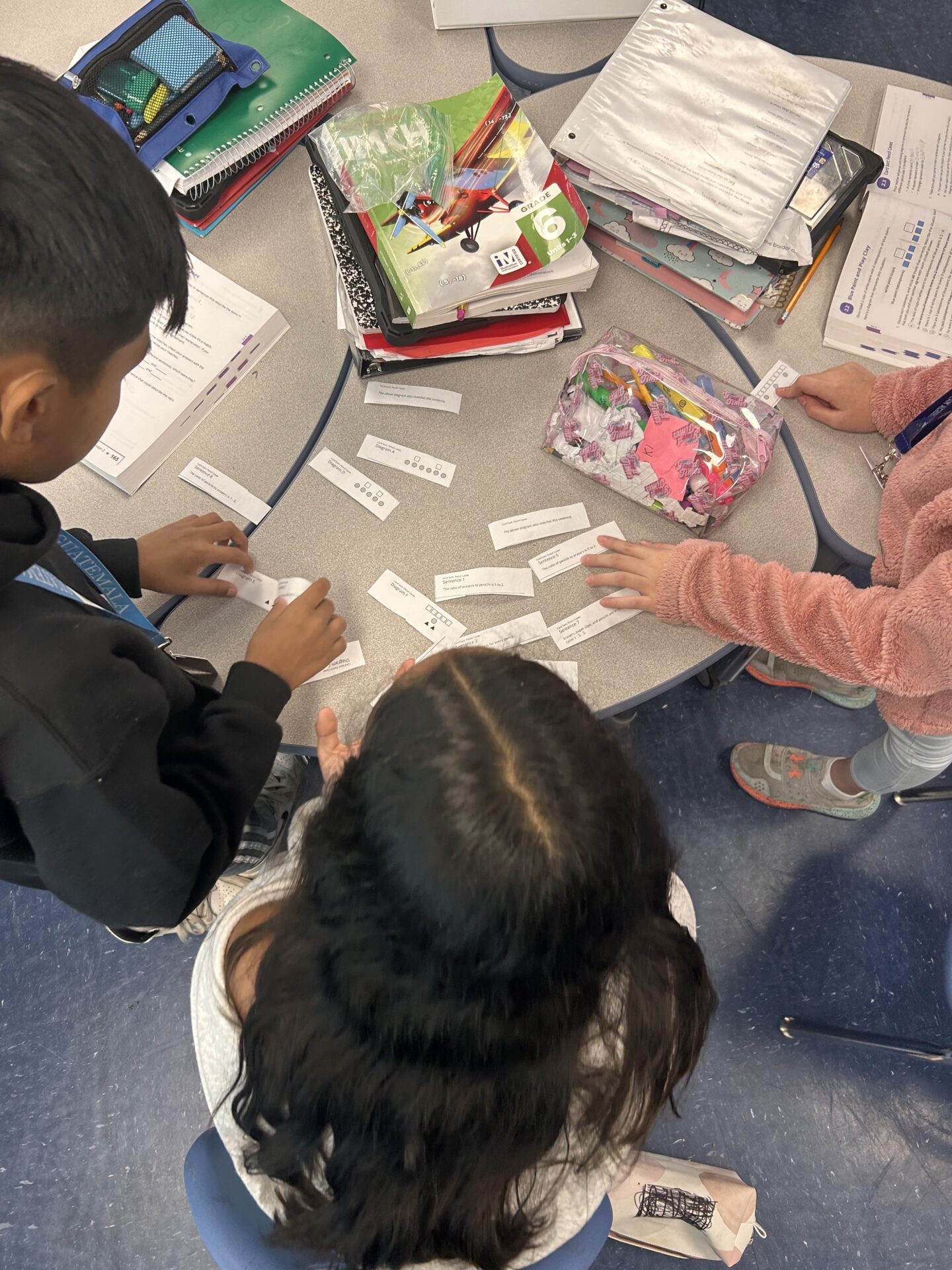 three children working together at a round table on what appears to be a math activity. They are arranging small slips of paper with printed numbers, diagrams, or math problems.