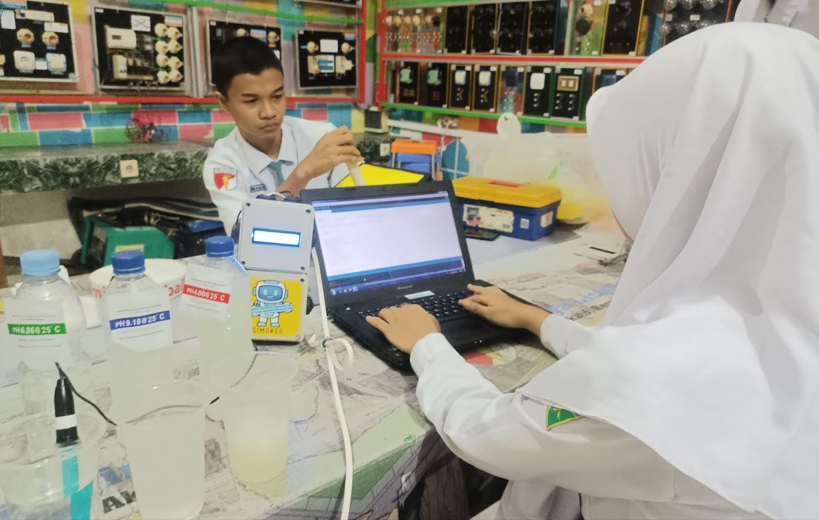 Two students sitting across from each other at a desk, one working on an open laptop.
