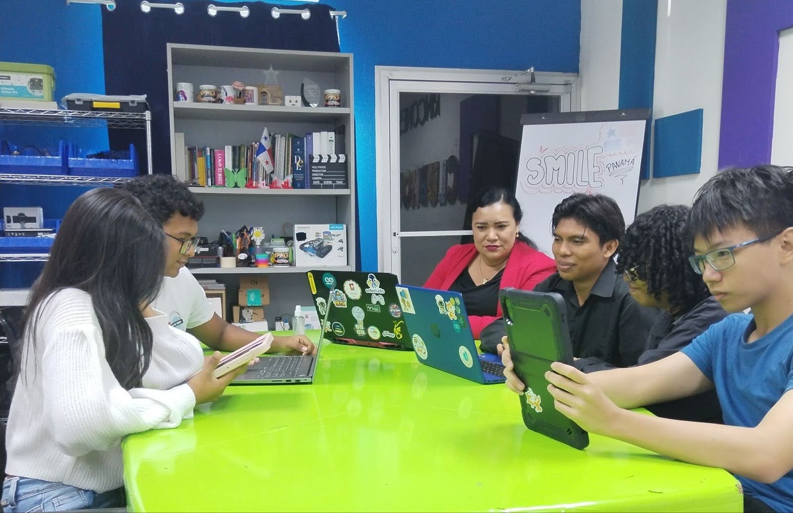 Five students and one educator seated around a desk, working on open laptops. 