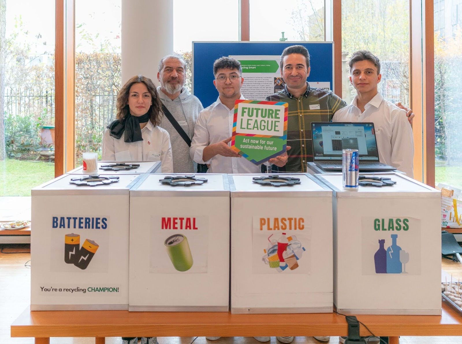 Students and teachers standing in front of an exhibit display featuring posters with the words Batteries, Metal, Plastic, and Glass, while holding a sign with the words Future League: Act Now for a Sustainable Planet.