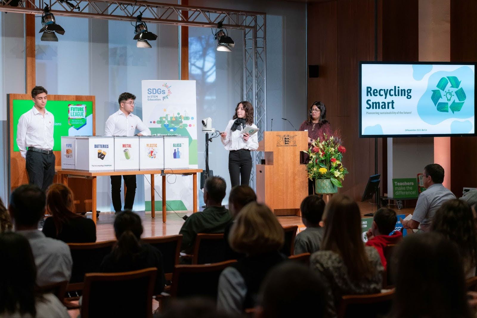 Students standing on a stage in front of an exhibit display featuring posters with the words: Batteries, Metal, Plastic, and Glass. Two women standing next to a computer projector with the words Recycling Smart on the display.