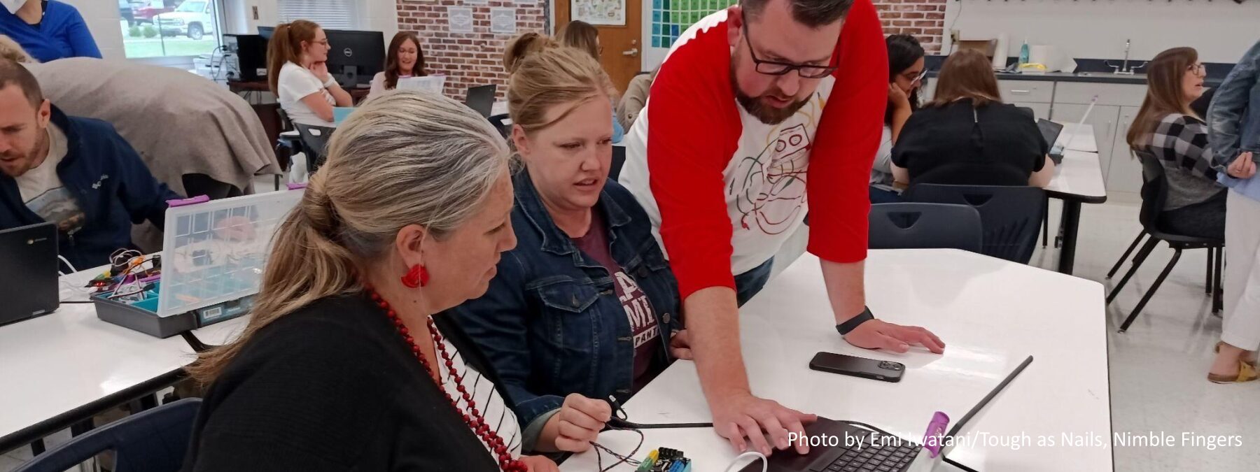 Three teachers at a table interact with a laptop and technology at a desk. Several other teachers organized in clusters are in the background.