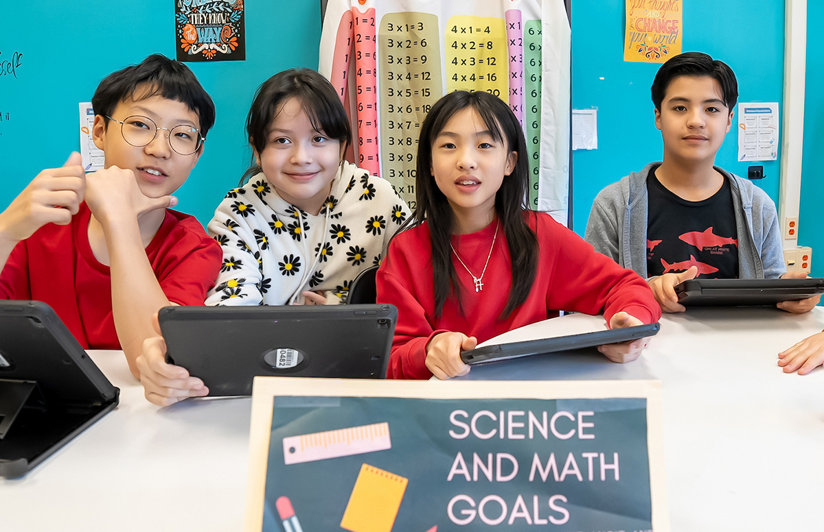 Four students at a classroom table with tablets, behind a sign reading "Science and Math Goals."