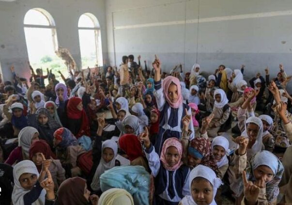 A large group of students sit in a classroom with some raising a pointed finger toward the ceiling