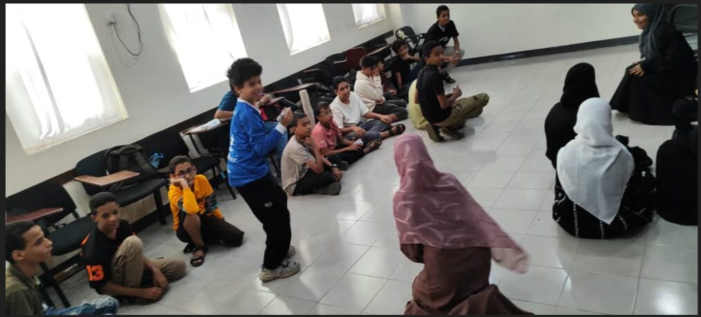 Students are seated on a classroom floor while two play in the center