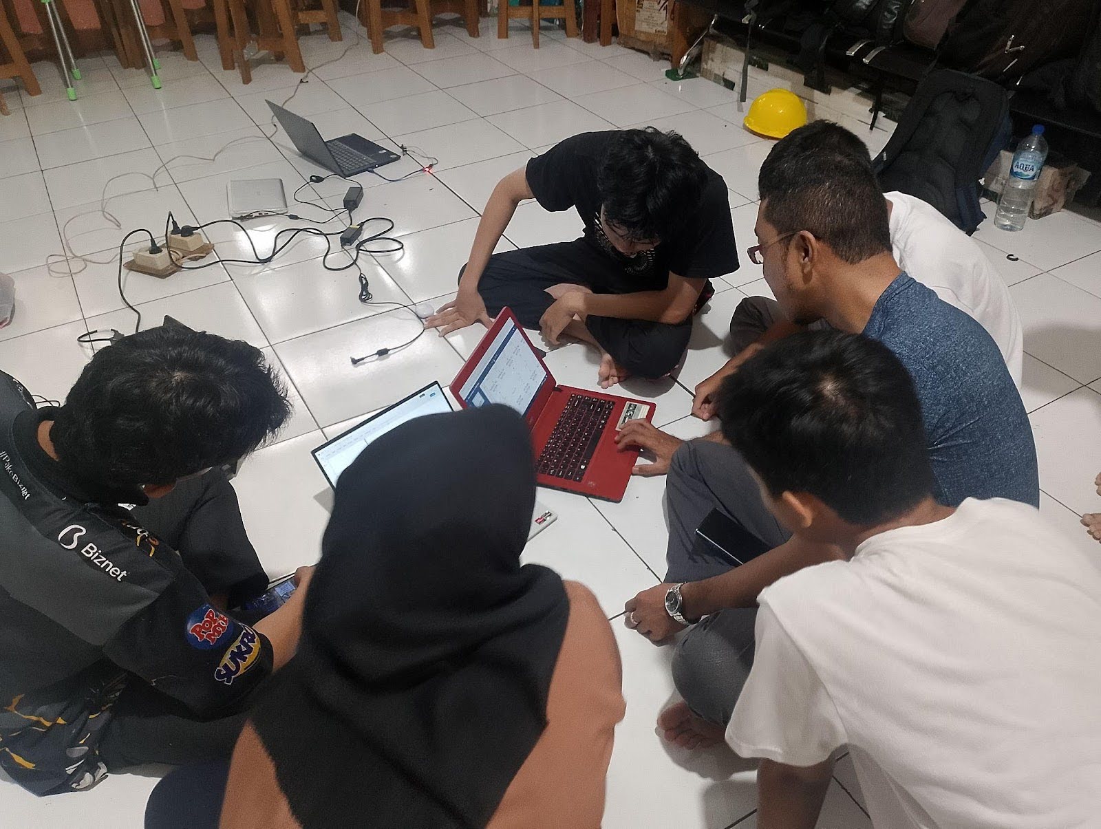 Four students huddle around a laptop and devices while sitting on the floor