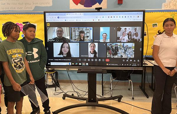Students pose for a picture next to a large TV screen with a Zoom meeting display