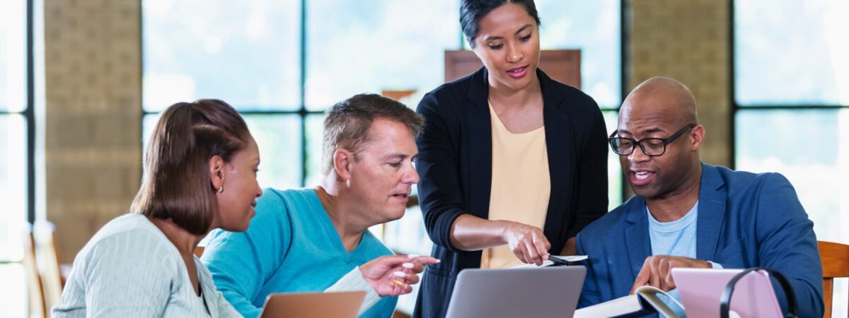 An image of a group of educators discussing and looking at each other's laptops. One educator in the center is standing while the others are sitting at a table.