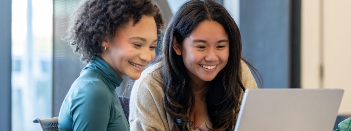 Two young girls smiling while collaborating on a laptop.