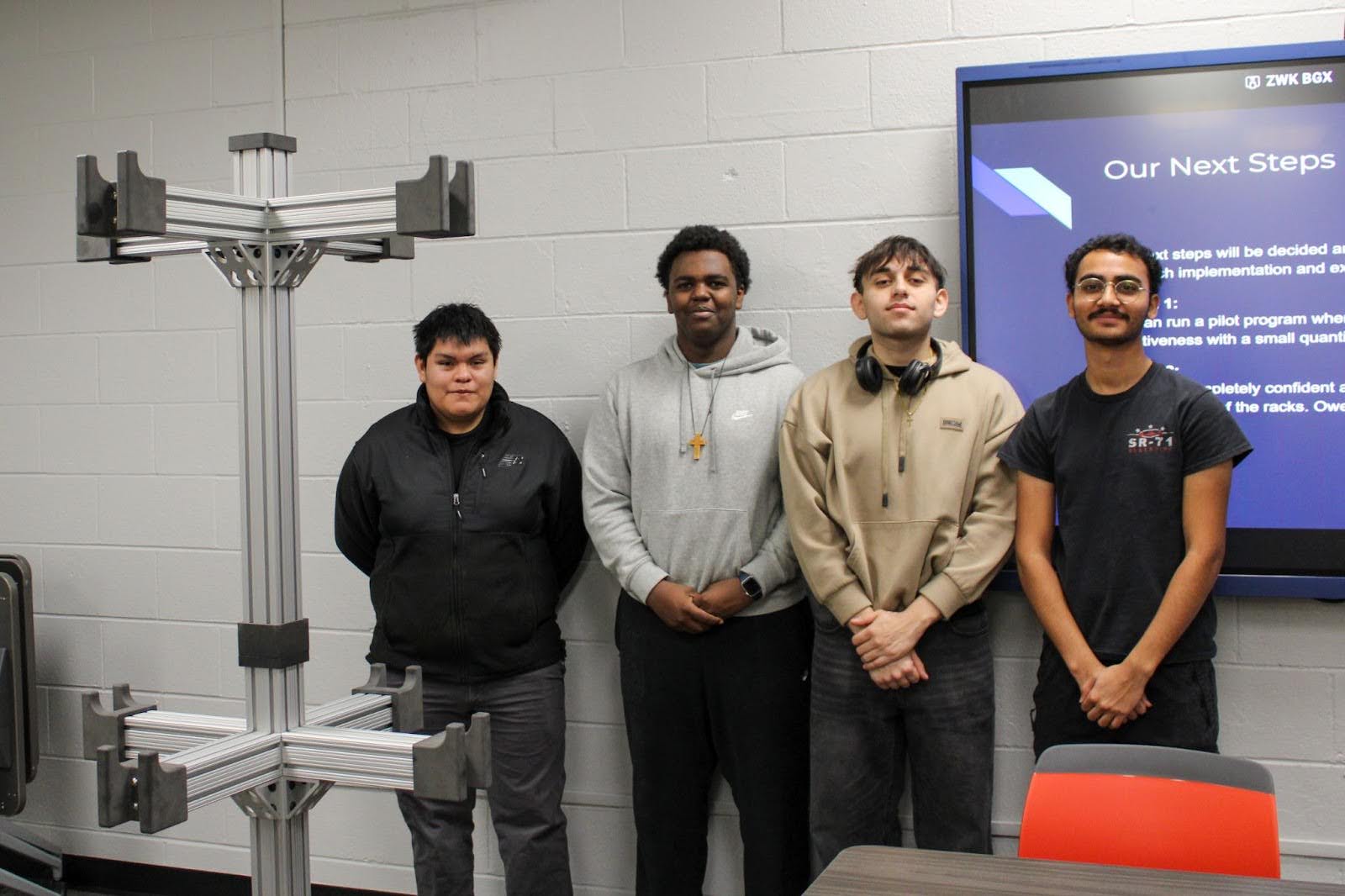 A group of four students standing and posing alongside a classroom project presentation.