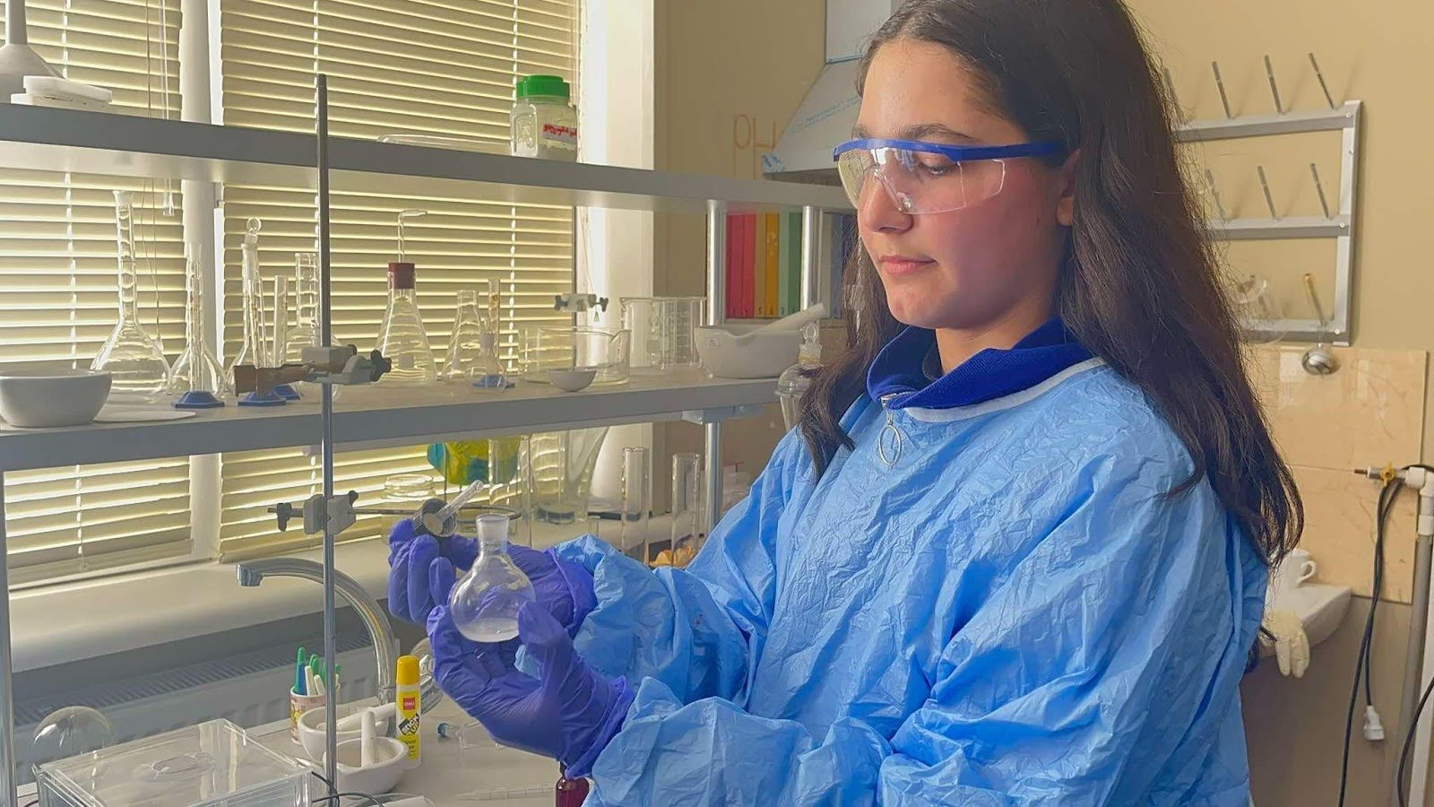 A student in a chemical lab wearing lab equipment such as gloves, goggles, and blue coat holding a glass mixer.