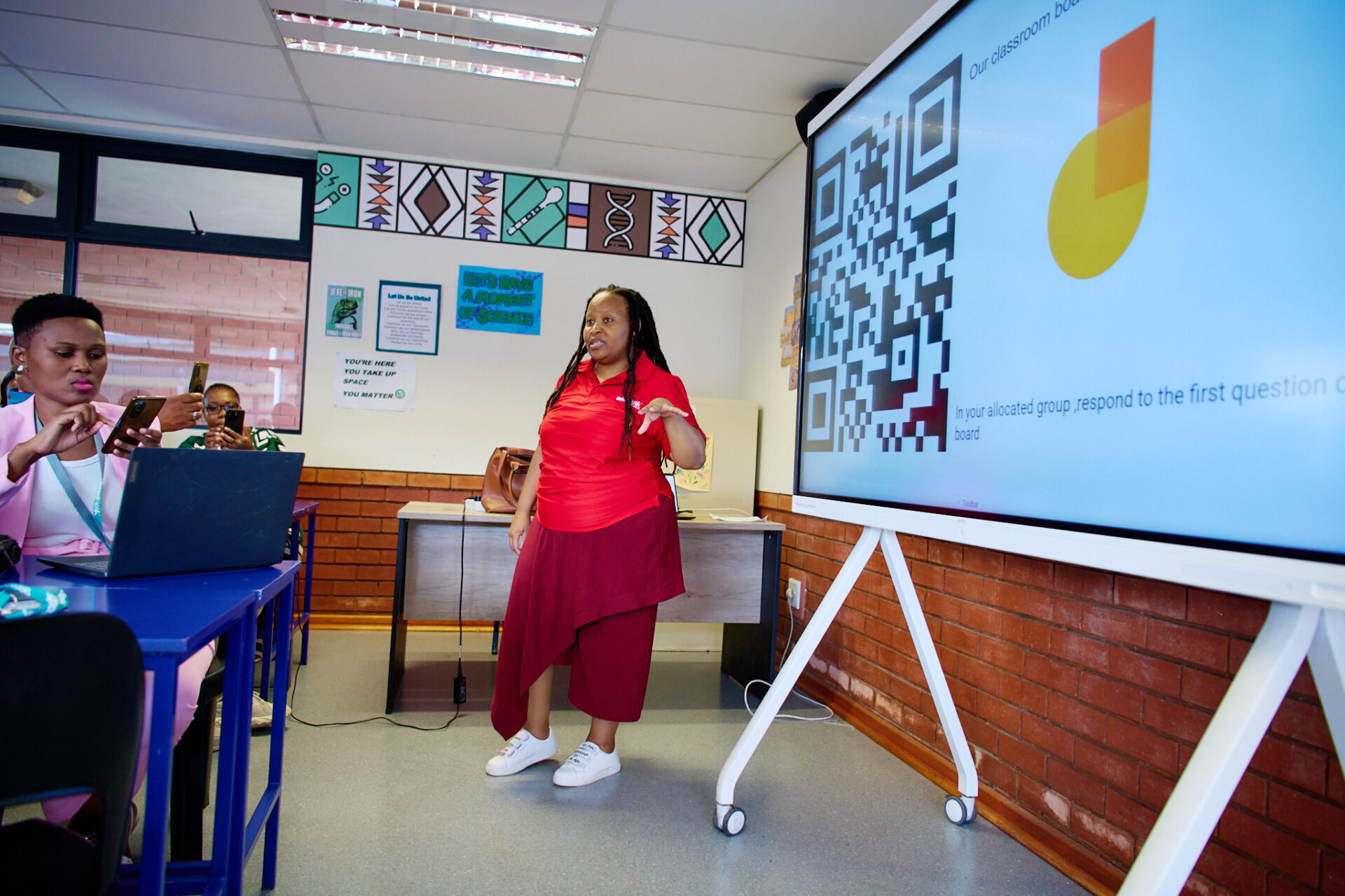 An instructor leads a classroom session using a digital whiteboard displaying a QR code, while participants scan the code with their phones and laptops to join the activity.