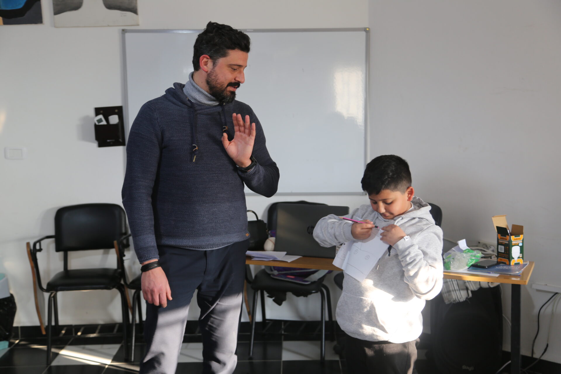 Young boy pointing at a piece of paper that he is holding against his chest while an adult facilitator looks on.