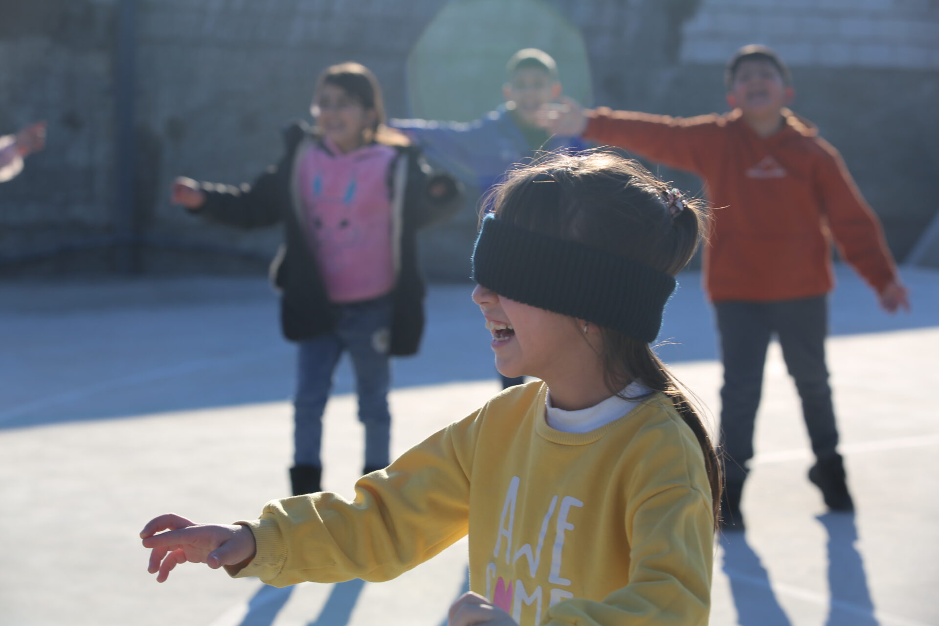 Young child laughing while wearing a blindfold, with other children in the background speaking and directing her.