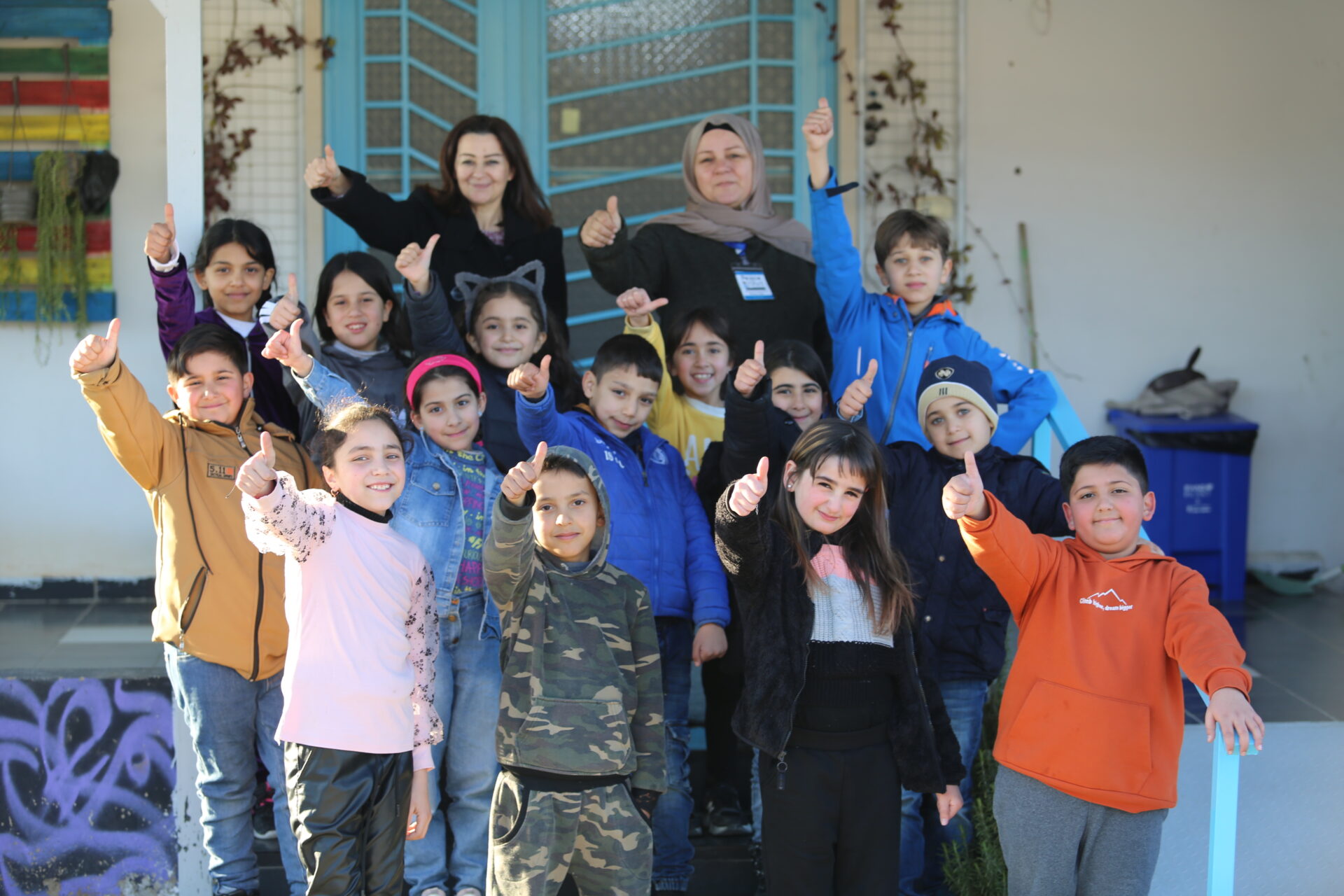 Group of 14 children and 2 adults smiling and giving a thumb’s up to the camera.