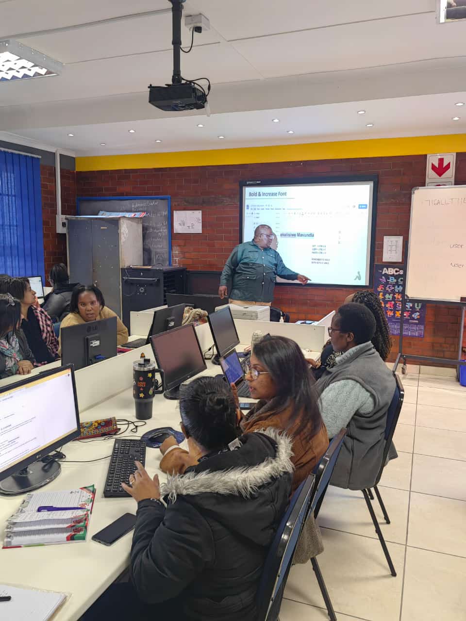 A group of educators in a computer lab participate in a training session, as an instructor at the front points to a projected screen with digital learning content.