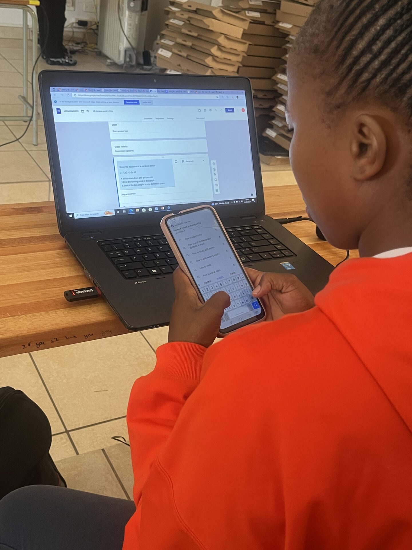 An educator uses a smartphone while working on a laptop displaying a digital class activity form.
