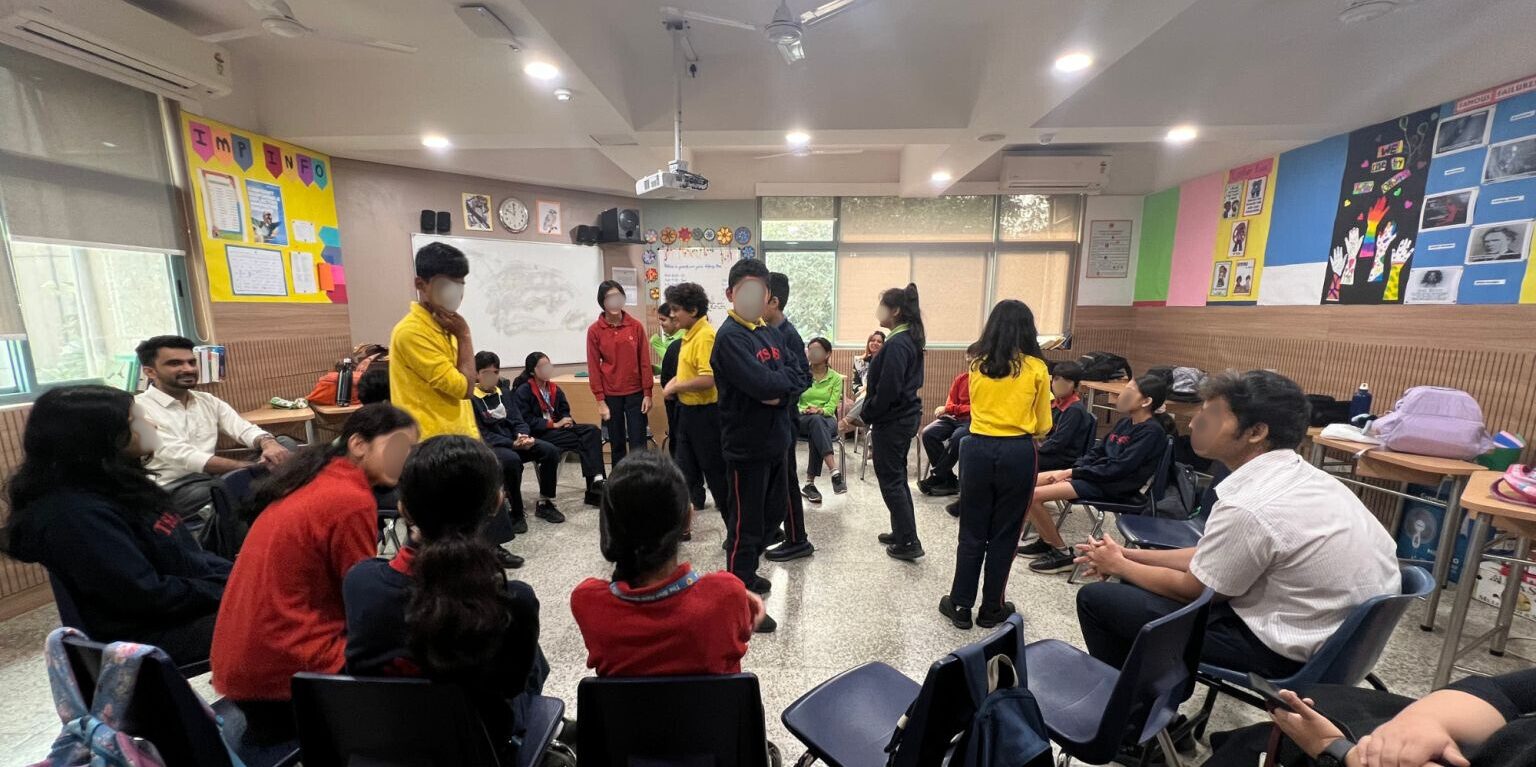 Students participate in a classroom discussion circle, with several standing in the center while others sit and observe.