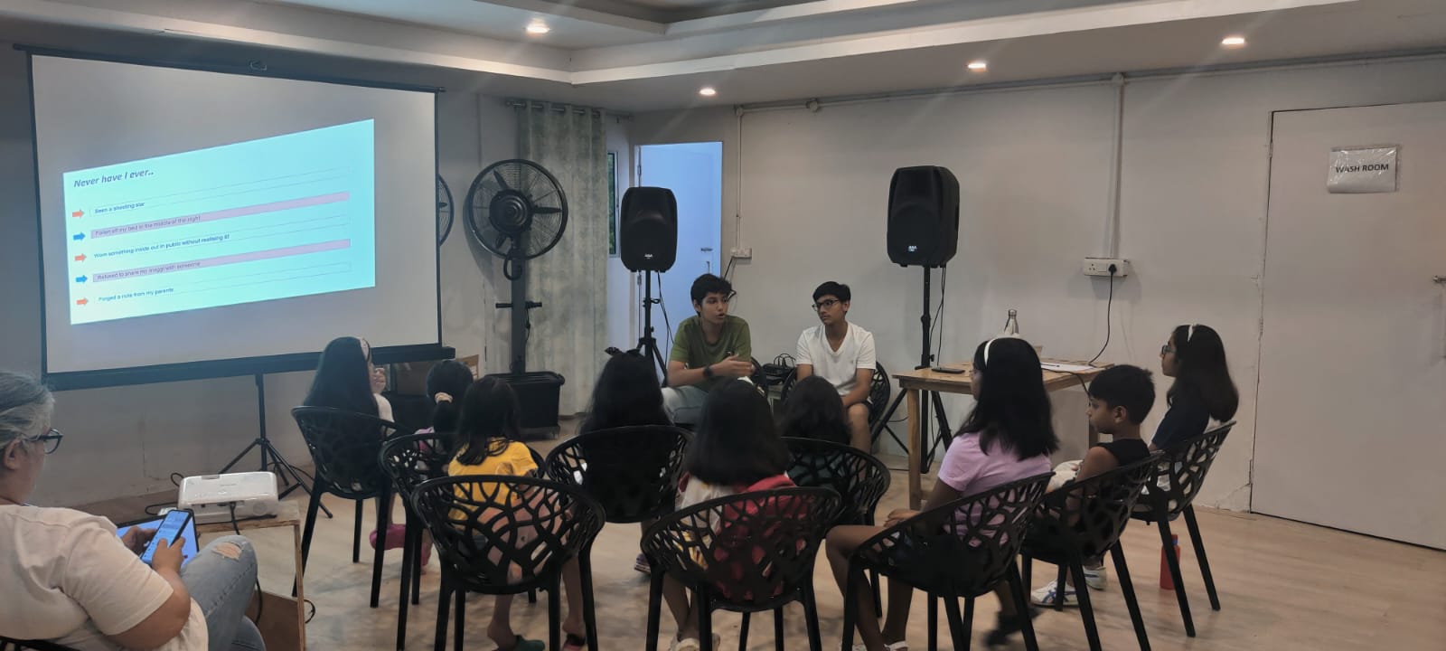 Students sit in a circle while two teens facilitate a discussion beside a projected slide.