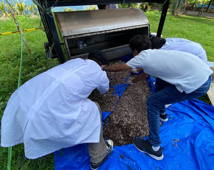 Students wearing protective coats gather composted food waste on a tarp beneath a composting machine.