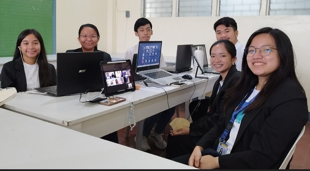 Students seated at a table with laptops and a tablet participate in a virtual meeting.