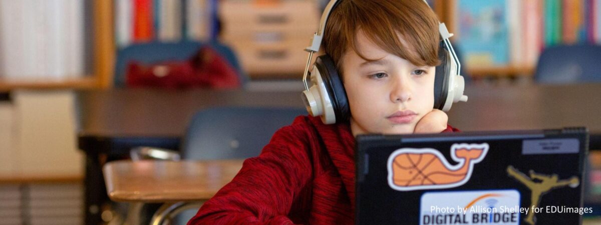 A middle school aged boy with headphones is seated at a desk with a laptop watching it.