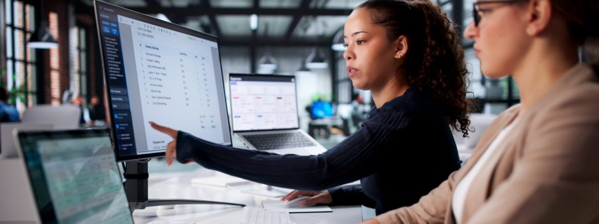 A professional points at a pricing document on a monitor while explaining it to a colleague in an office.