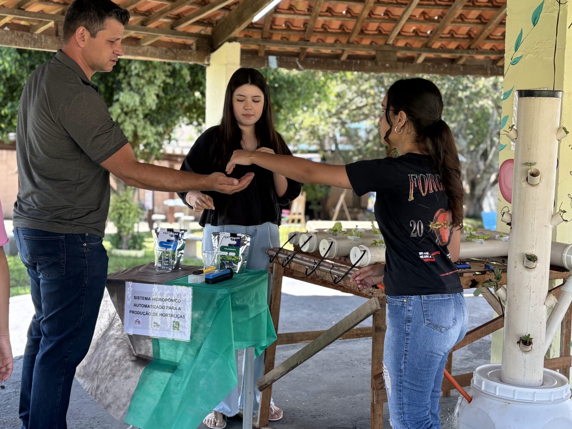 Two students and one teacher stand in a circle around a hydroponics system, with one student placing seeds in the teacher's hand