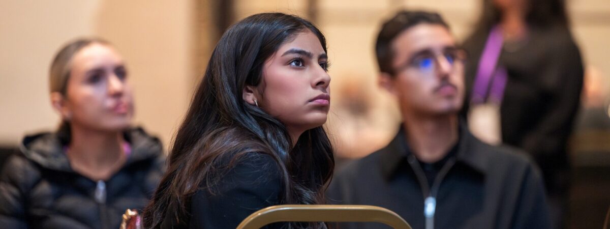 A photo of a student with long hair sitting in a chair looking up at a speaker on a stage.