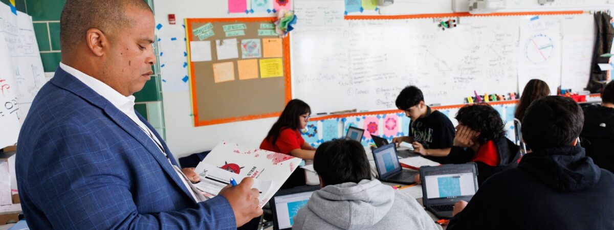 A man in a suit stands with a clipboard and pen in front of a classroom students using laptops.