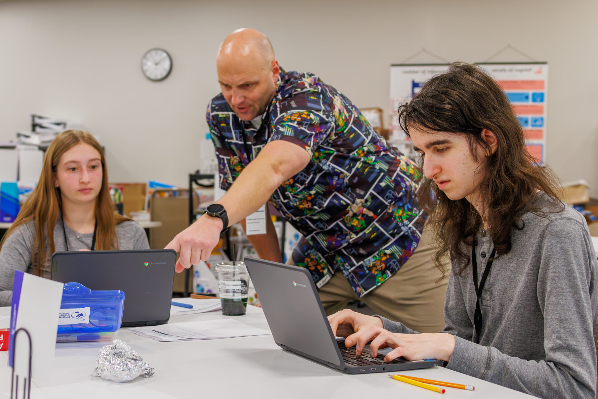 Two students are seated at a table with laptops in front of them while a teacher points at one of the student's screen