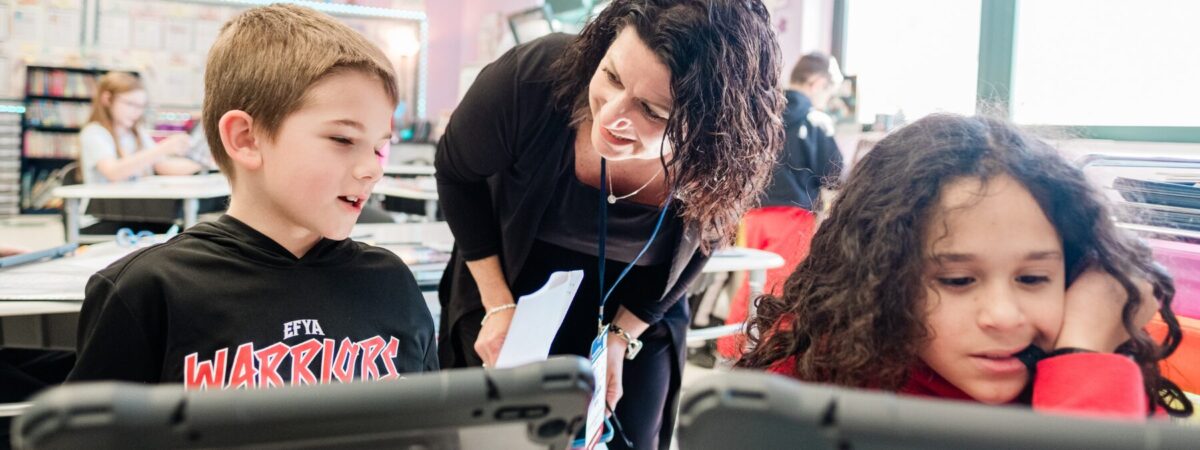 Adult woman interacting with younger male student, next to female student, with both students in front of tablets in classroom setting