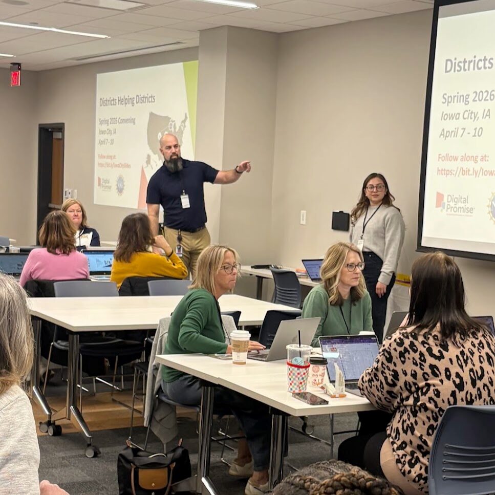 A man wearing a polo stands in the middle of a classroom of adults, pointing at a presentation on a white screen on a wall. 