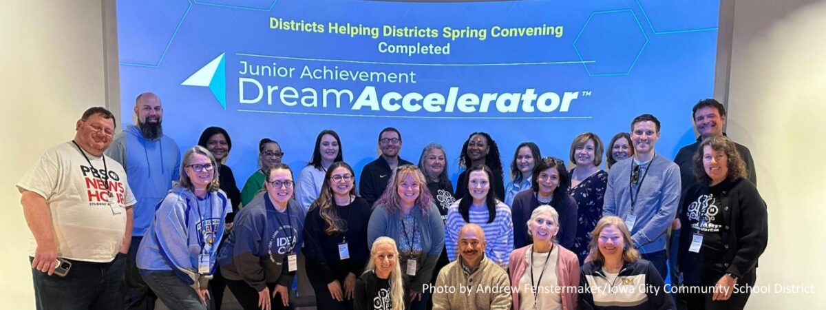 A group of about 30 people are standing on a stage in front of a screen that reads: Districts Helping Districts Spring Convening Completed, Junior Achievement Dream Accelerator