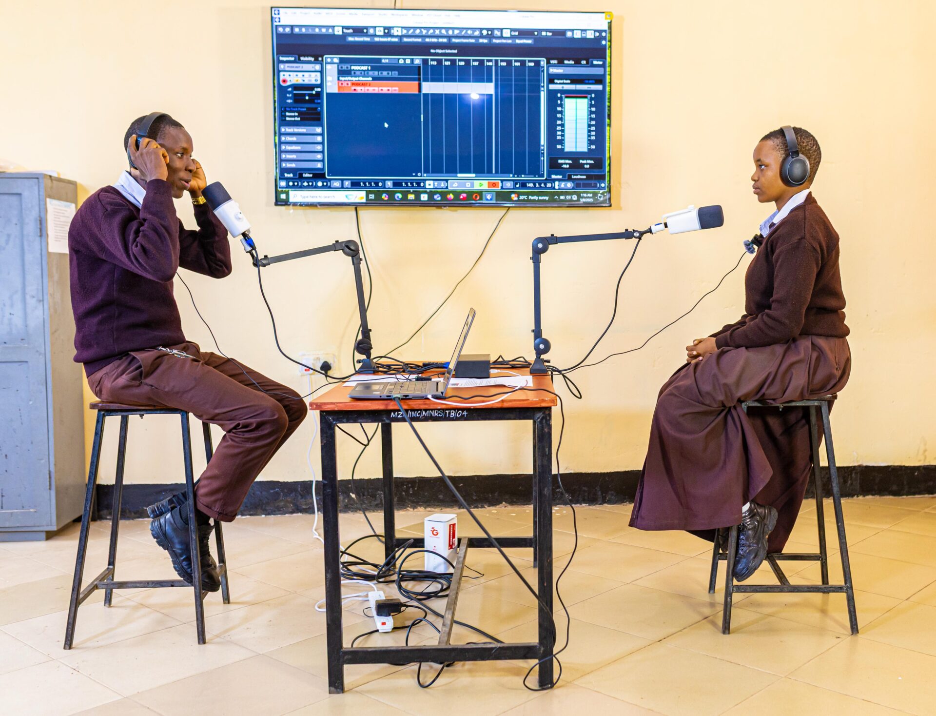 Wisdom and Neema sit facing each other while recording a podcast in a classroom.