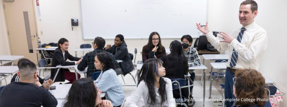 Multiethnic group of students talking with instructor in a classroom with other groups on a college campus.